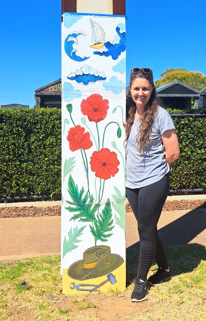 Artist Joanna Hubbard stands next to stobie pole which displays her artwork showing a soldier's hat and tools at the base, red poppies in the middle and a sailling ship in the clouds above.