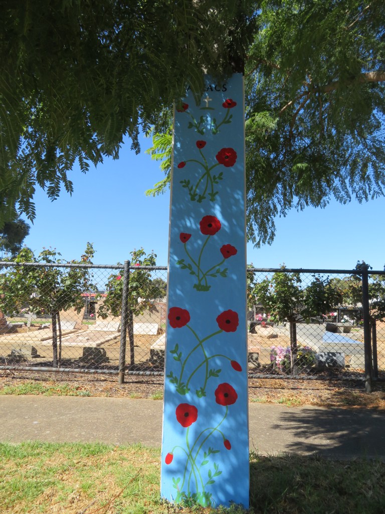 A stobie pole art of red poppies on light blue background. Cheltenham Cemetery in background.